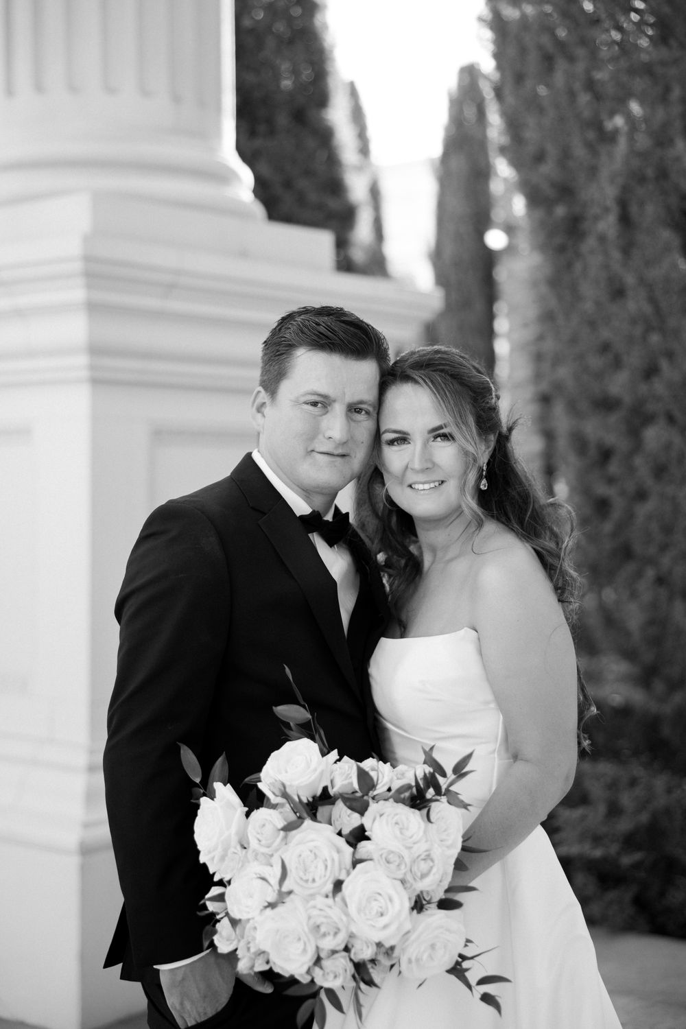 Black and white portrait of smiling bride and groom posing with white rose bouquet near elegant columns and cypress trees.
