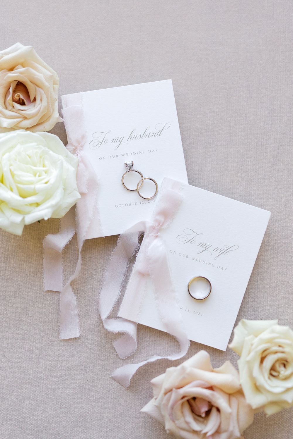 Elegant wedding flat lay with gold rings, vow cards, blush ribbon, and cream roses on a neutral background.