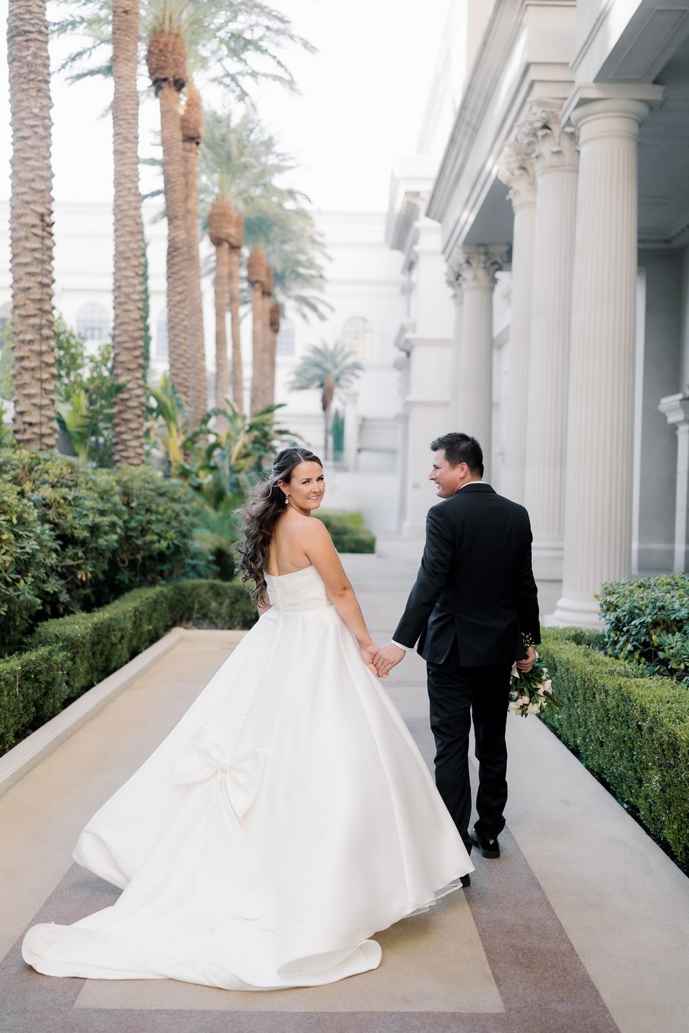 Bride in strapless white ballgown holds grooms hand walking past palm trees and white columns at elegant venue.