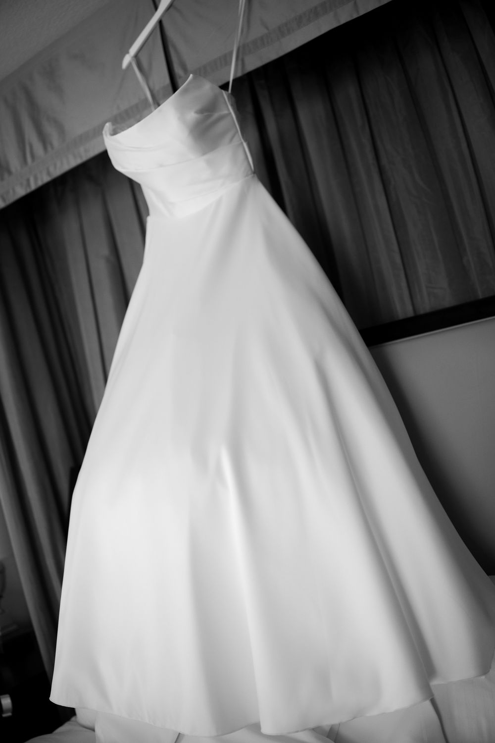 Black and white photo of a strapless ball gown wedding dress hanging on a hanger against draped curtains.