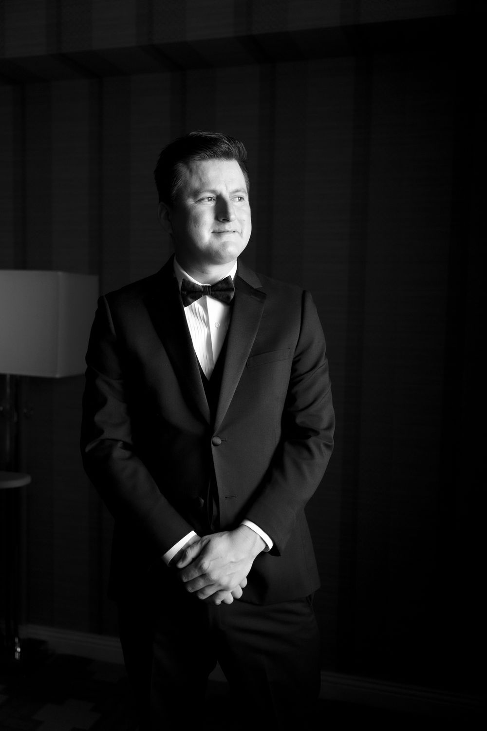 Groom in black tuxedo and bow tie poses confidently in dramatic black and white wedding portrait.