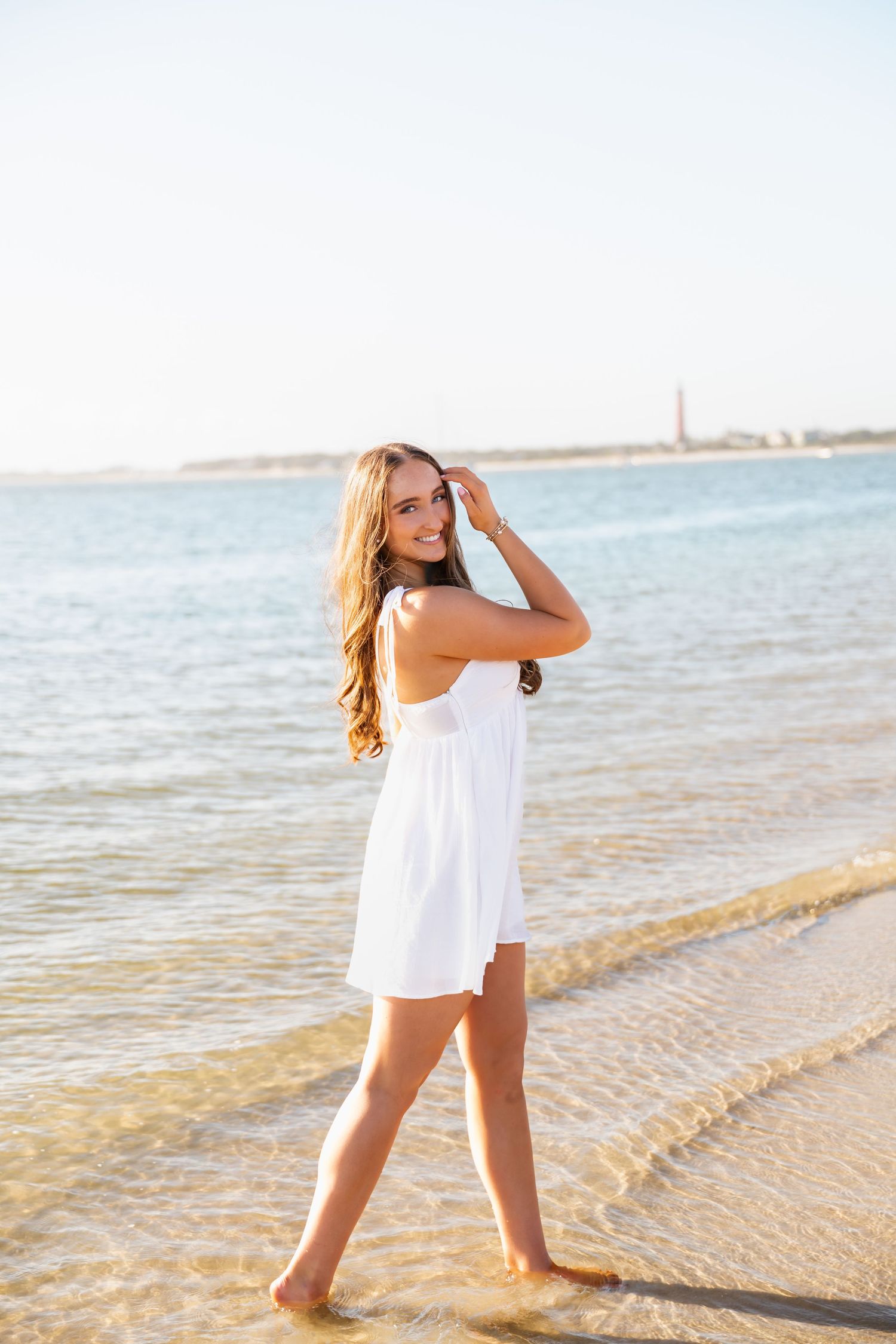 Young woman in white sundress walking barefoot on sunny beach shoreline with calm blue ocean waters behind her.