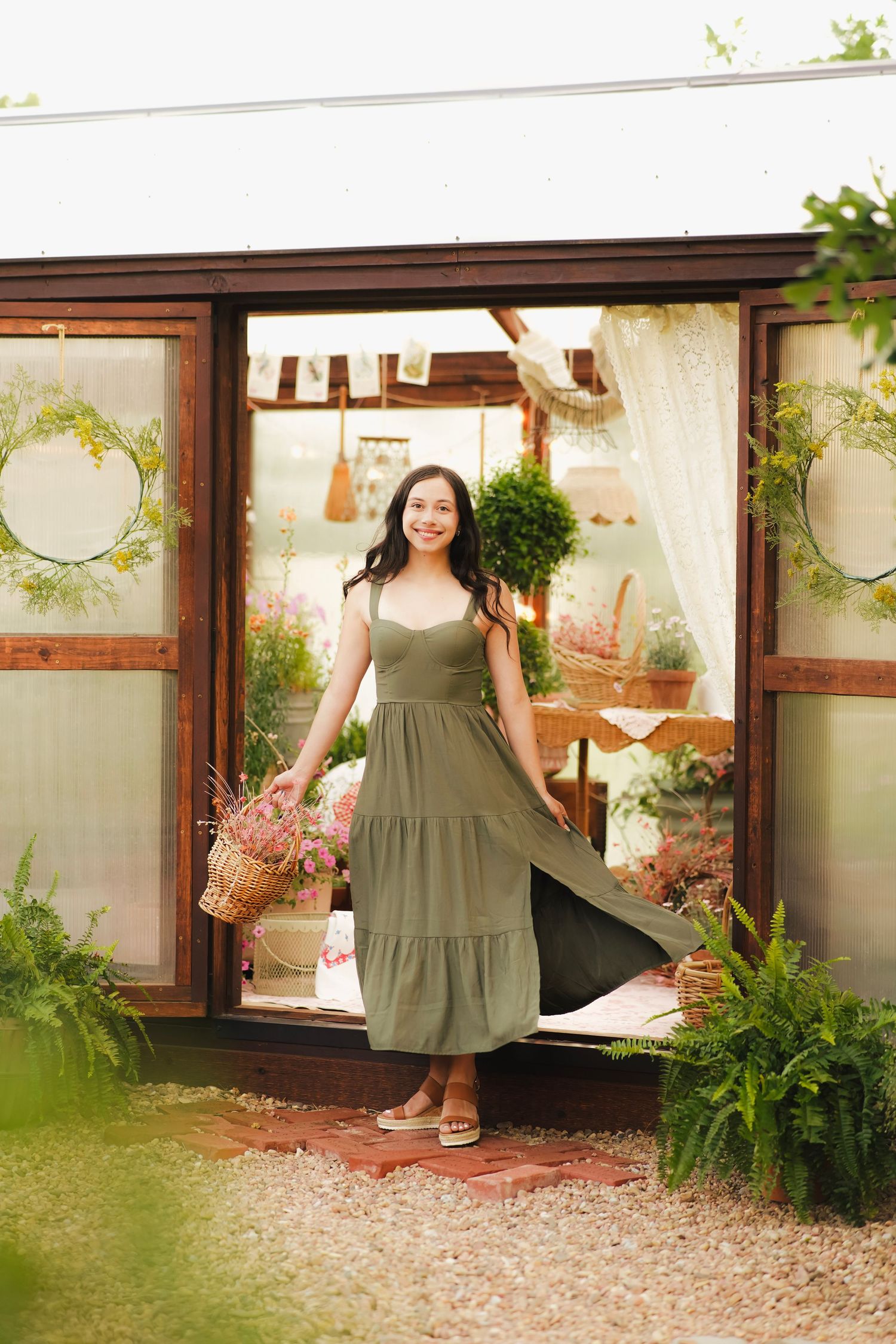 Smiling woman in olive green maxi dress holding flower basket standing in greenhouse doorway.