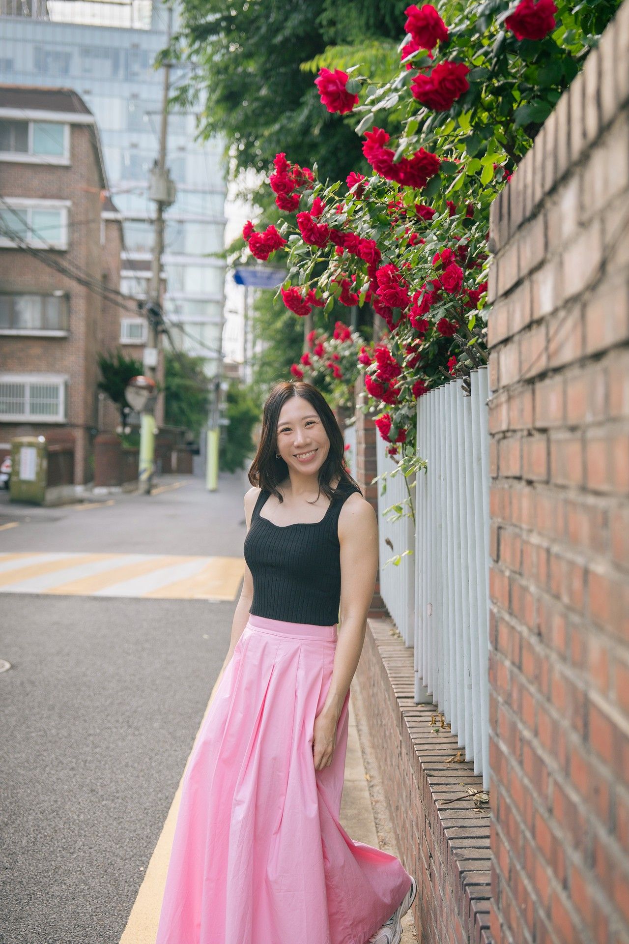 Woman in black top and pink skirt smiling beside a brick wall with red roses in an urban street setting.