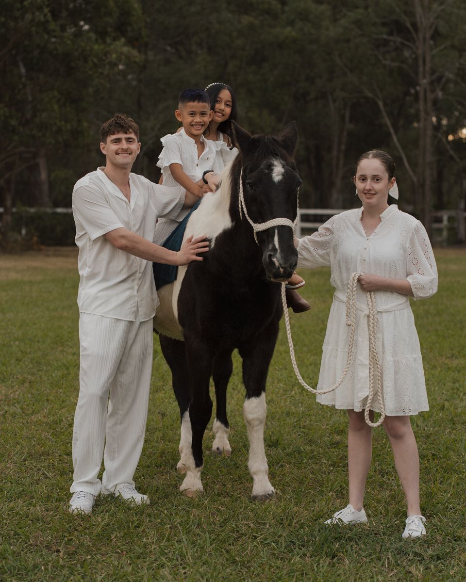 siblings in white outfits posing with a black and white horse on a green lawn with trees in background during family photoshoot with a horse