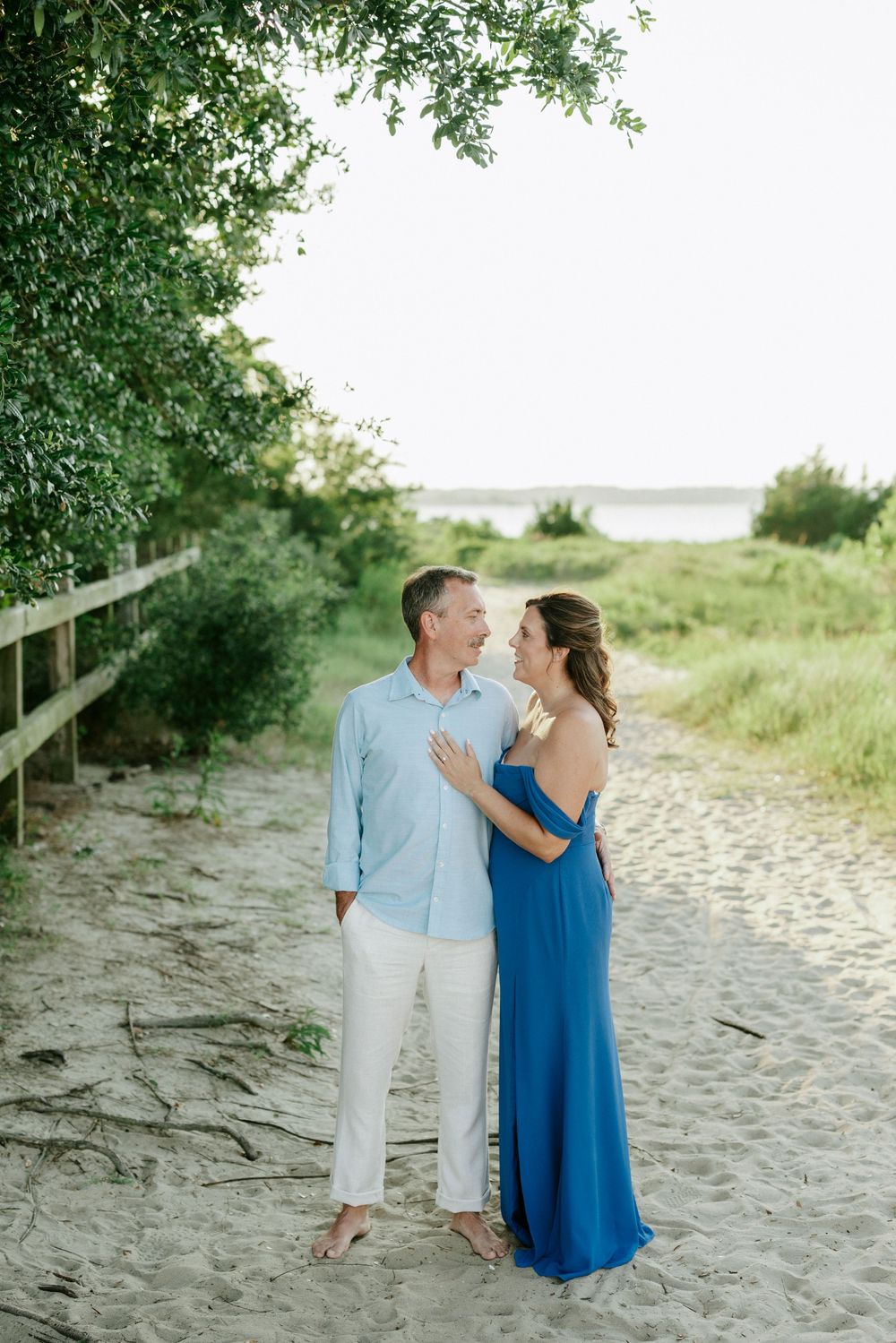 Couple in blue dress and light shirt embracing on sandy beach path surrounded by lush greenery.