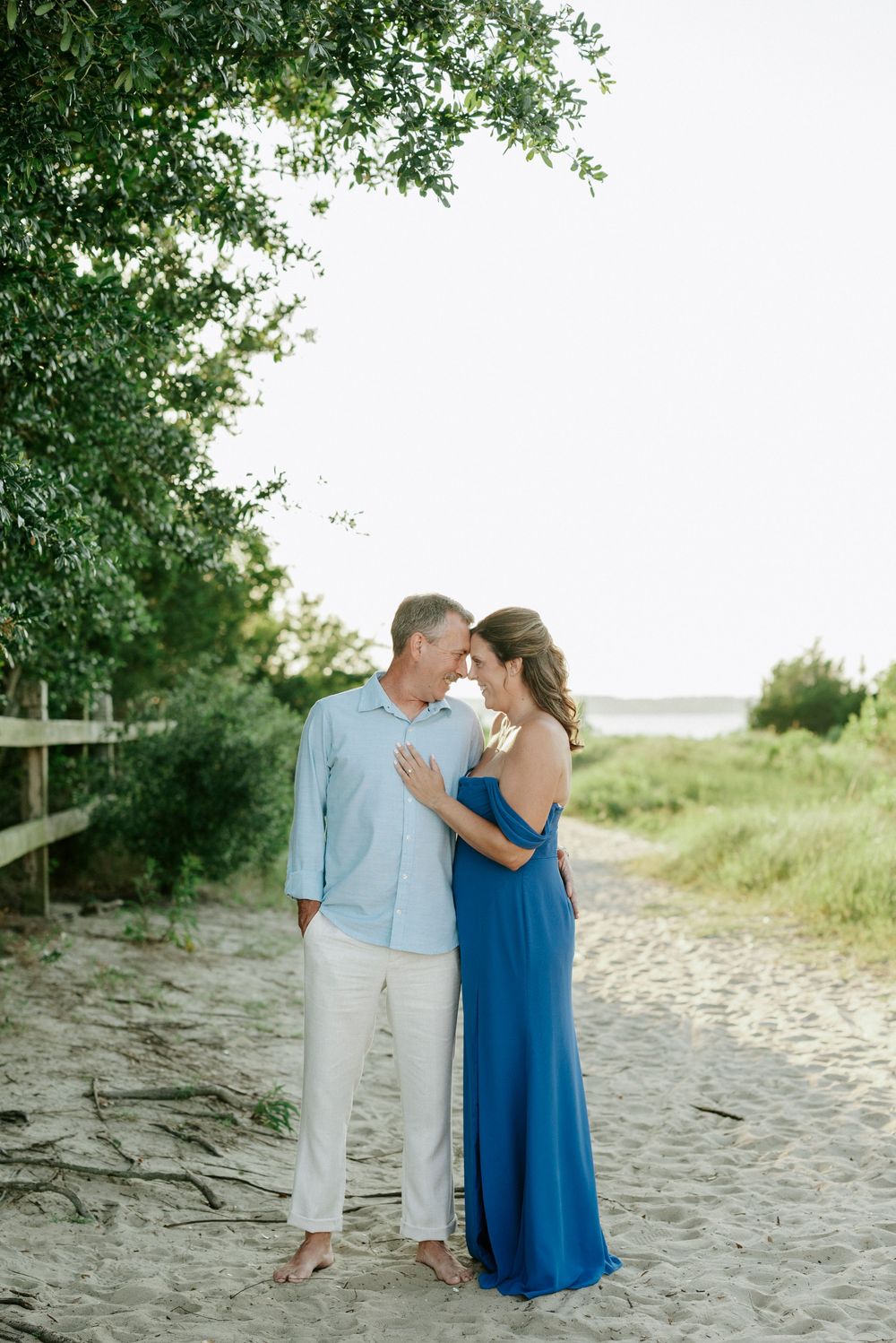 Couple sharing a romantic moment on a sandy beach path, woman in blue gown and man in light blue shirt.