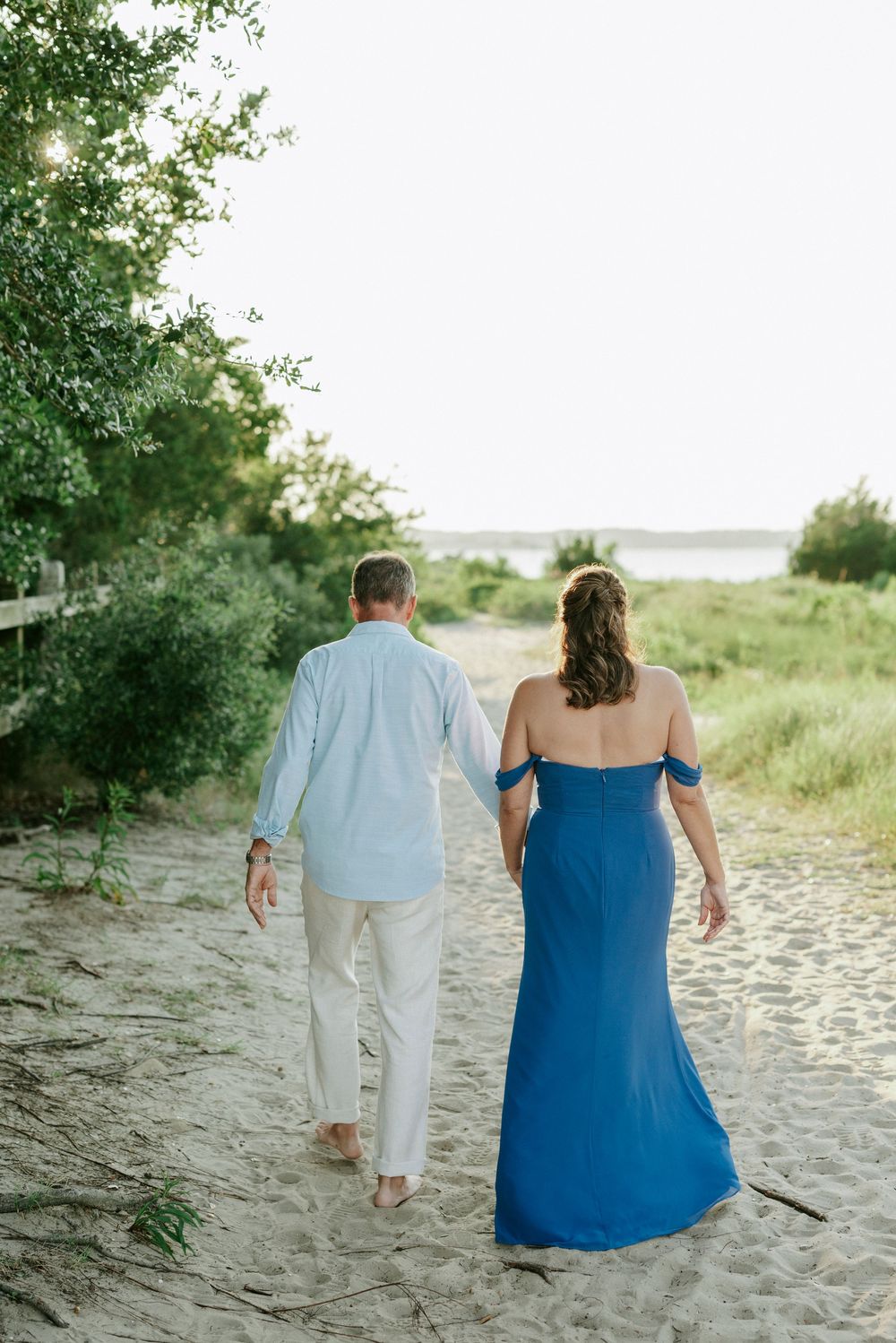 Couple walking hand-in-hand on sandy beach path, woman in blue off-shoulder gown, man in white linen outfit.