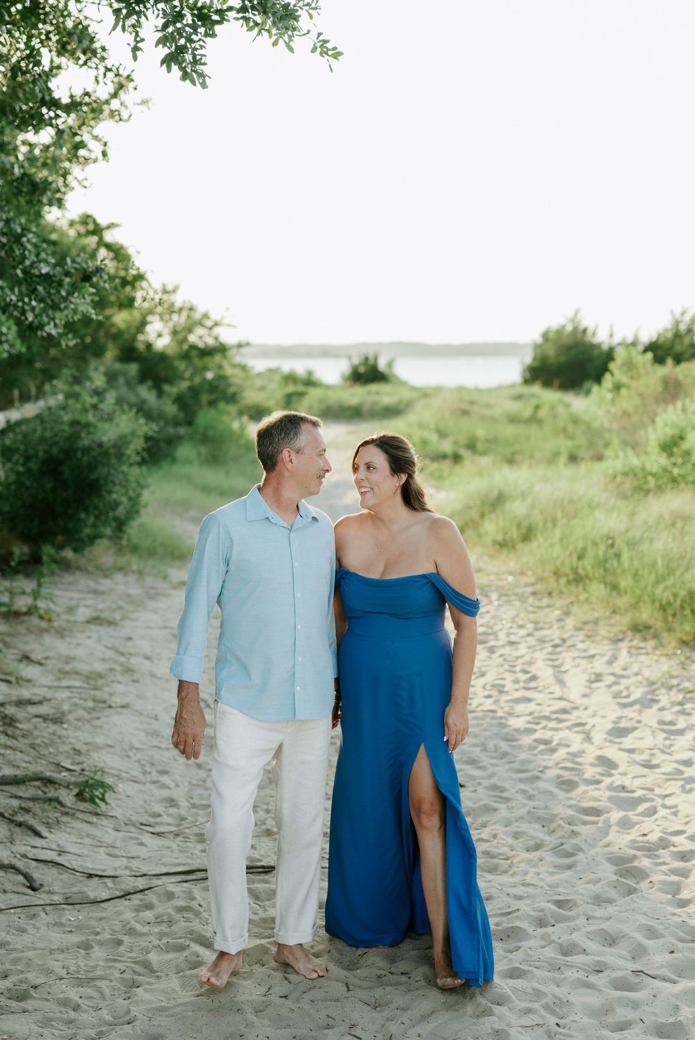 Couple walking hand-in-hand on sandy beach path, woman in blue off-shoulder gown, man in light blue shirt.