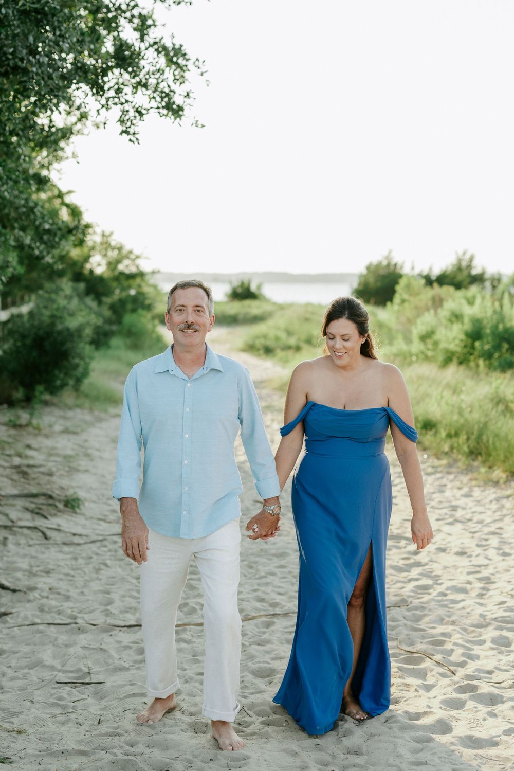 Happy couple walking hand-in-hand on a sandy beach path, woman in blue off-shoulder gown and man in white pants.