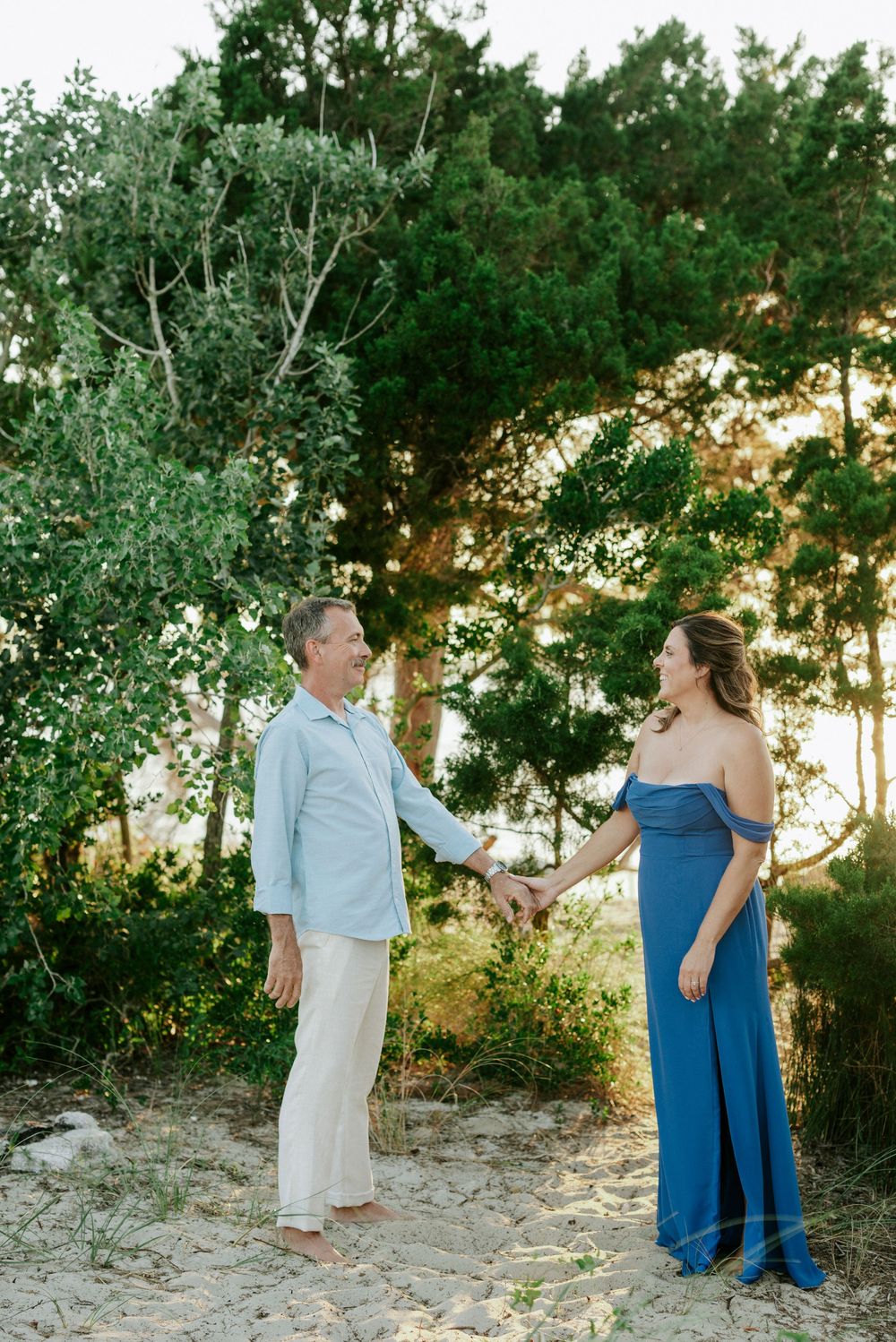 Couple holding hands and smiling on sandy path with tall green trees in warm sunlight.