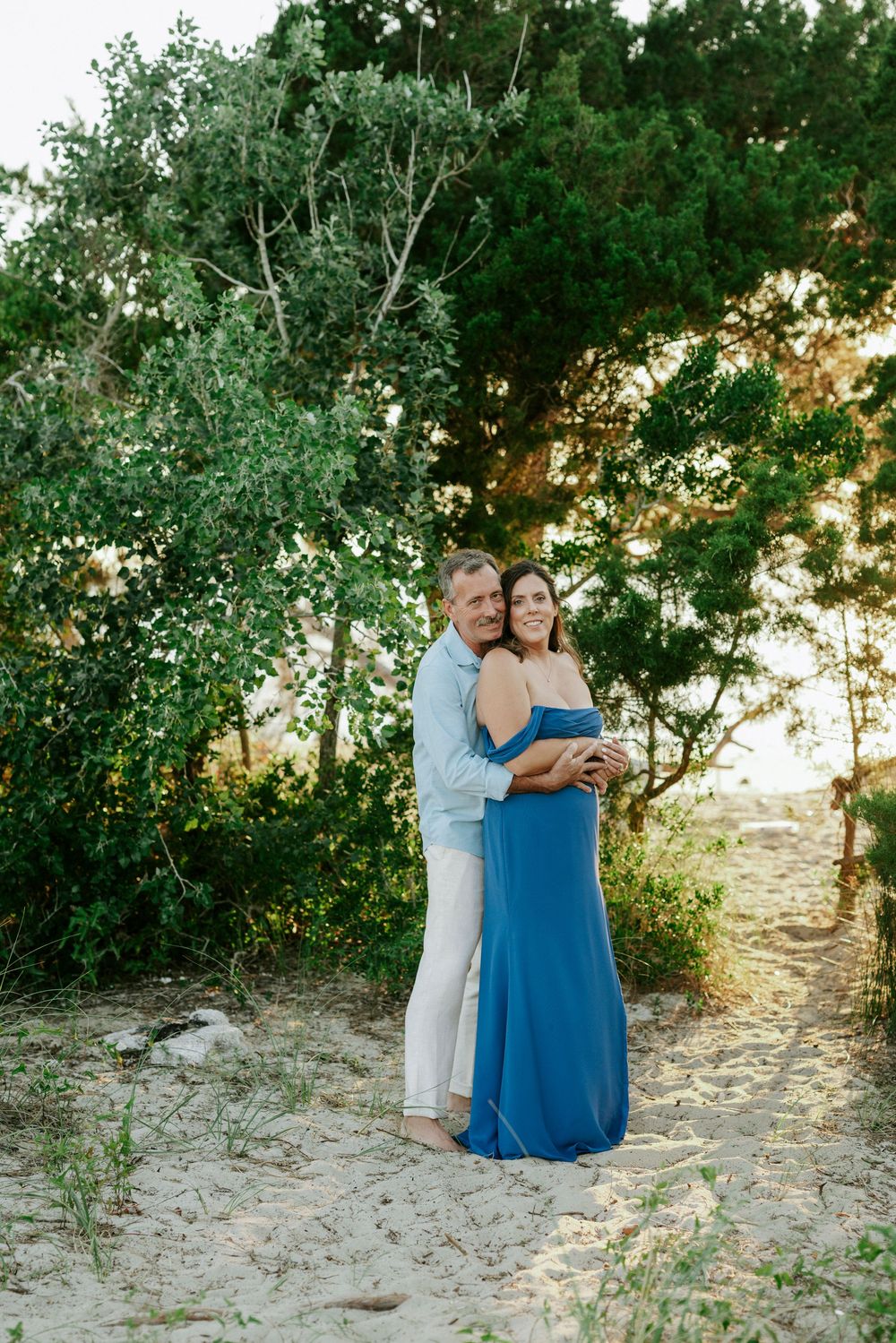 Man embracing woman in blue maxi dress on sandy path surrounded by lush green trees.