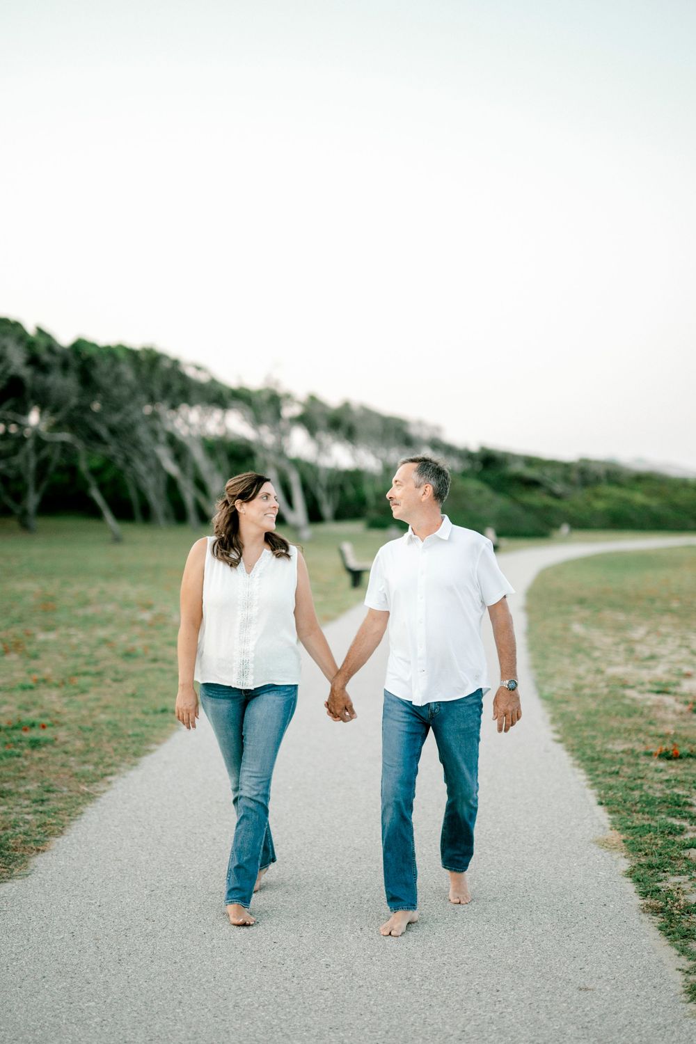 Couple in matching white shirts and jeans walking barefoot on a paved park path, smiling at each other.