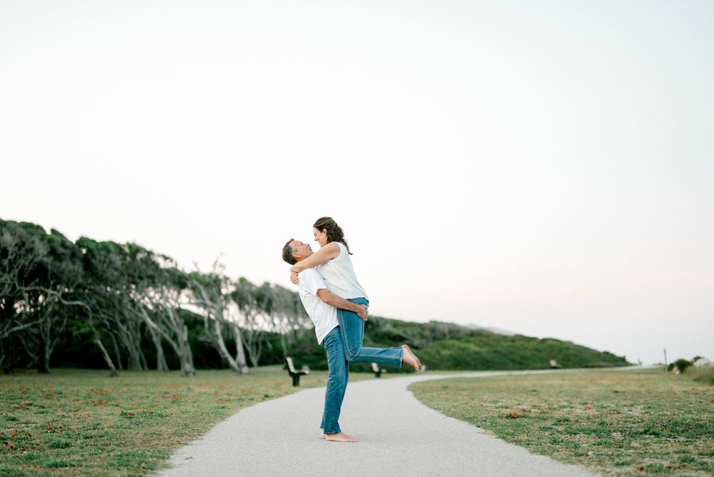 Man lifting woman in joyful embrace on scenic coastal pathway surrounded by lush greenery.