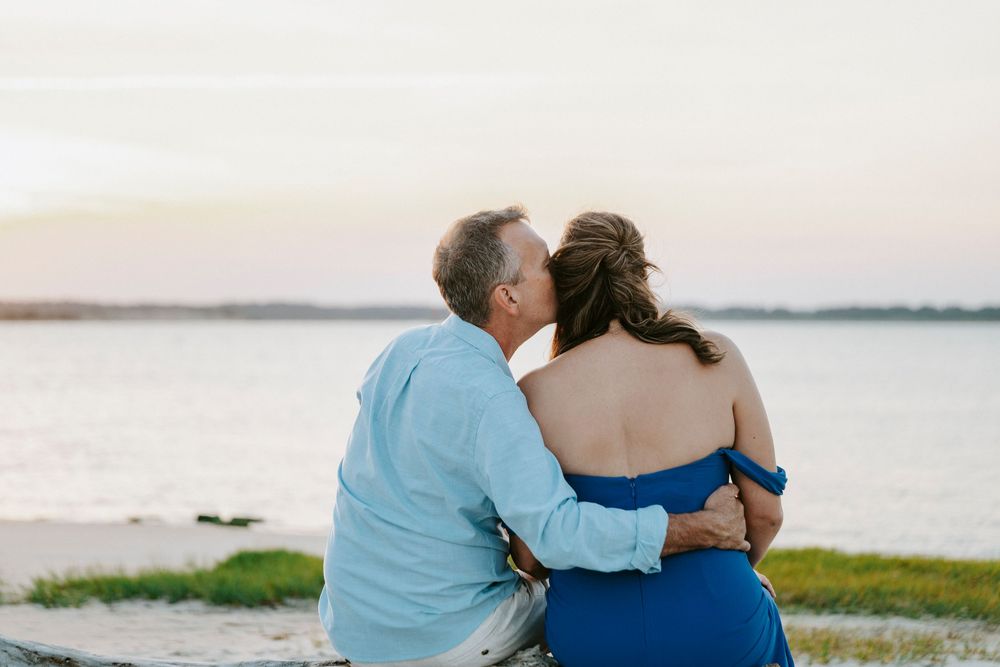 Couple sharing a romantic kiss at sunset by the water, man in blue shirt embracing woman in blue dress.
