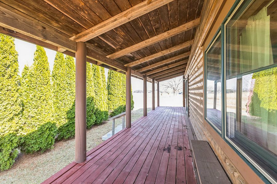 Covered wooden deck with rustic beam ceiling, red-stained floor, and lush green hedges.
