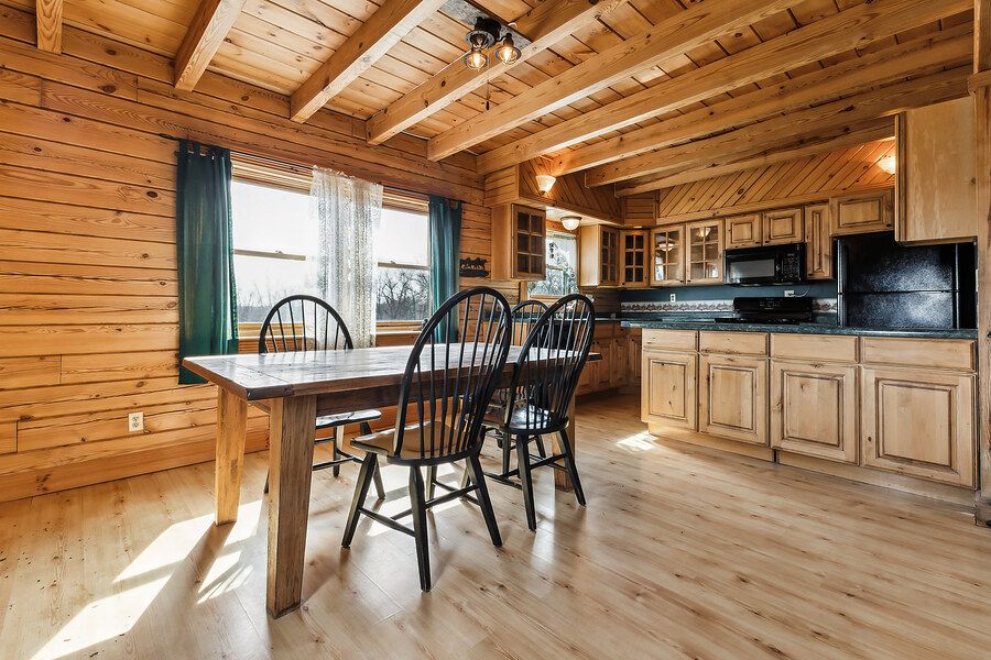 Bright log cabin dining area with wood beam ceiling, hardwood floors, and adjacent kitchen with oak cabinets.