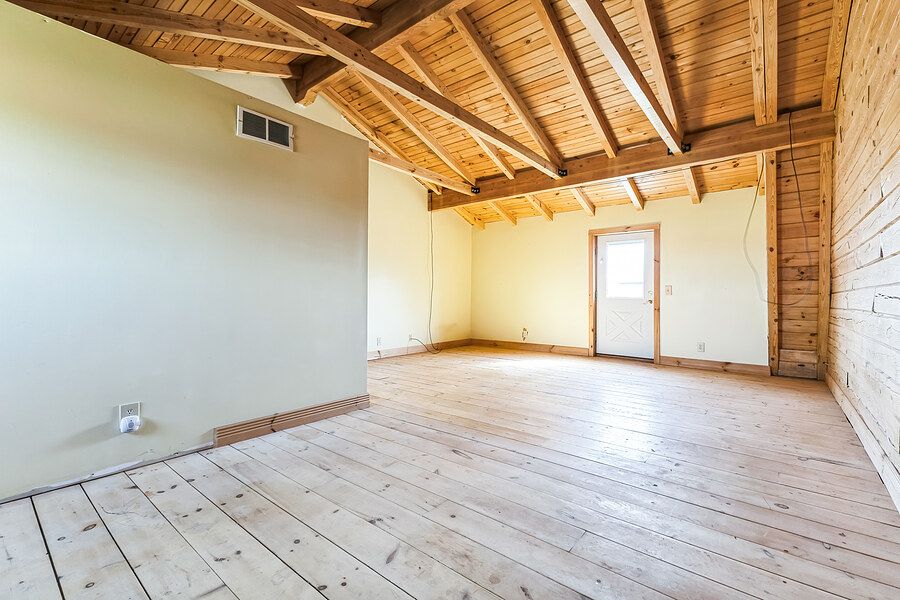 Spacious empty room featuring exposed wood beam ceiling, light hardwood floors, and a door with natural light.
