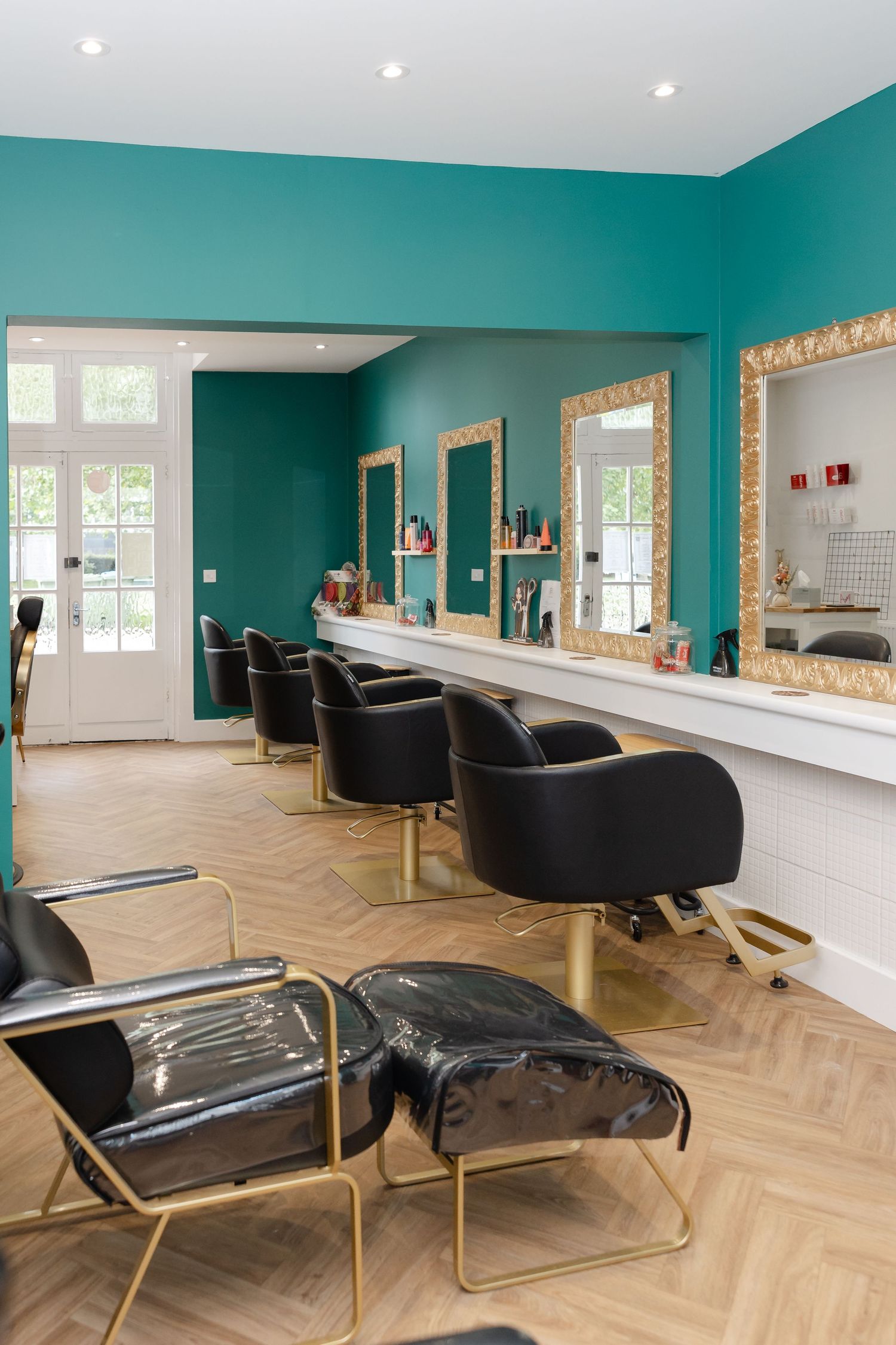 Modern hair salon with teal walls, black and gold styling chairs, mirrors, and herringbone wood floors.