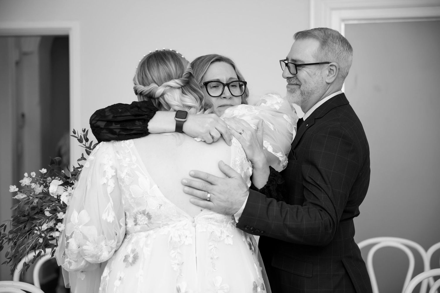 Black and white wedding photo of bride hugging guests while groom smiles warmly during indoor ceremony.