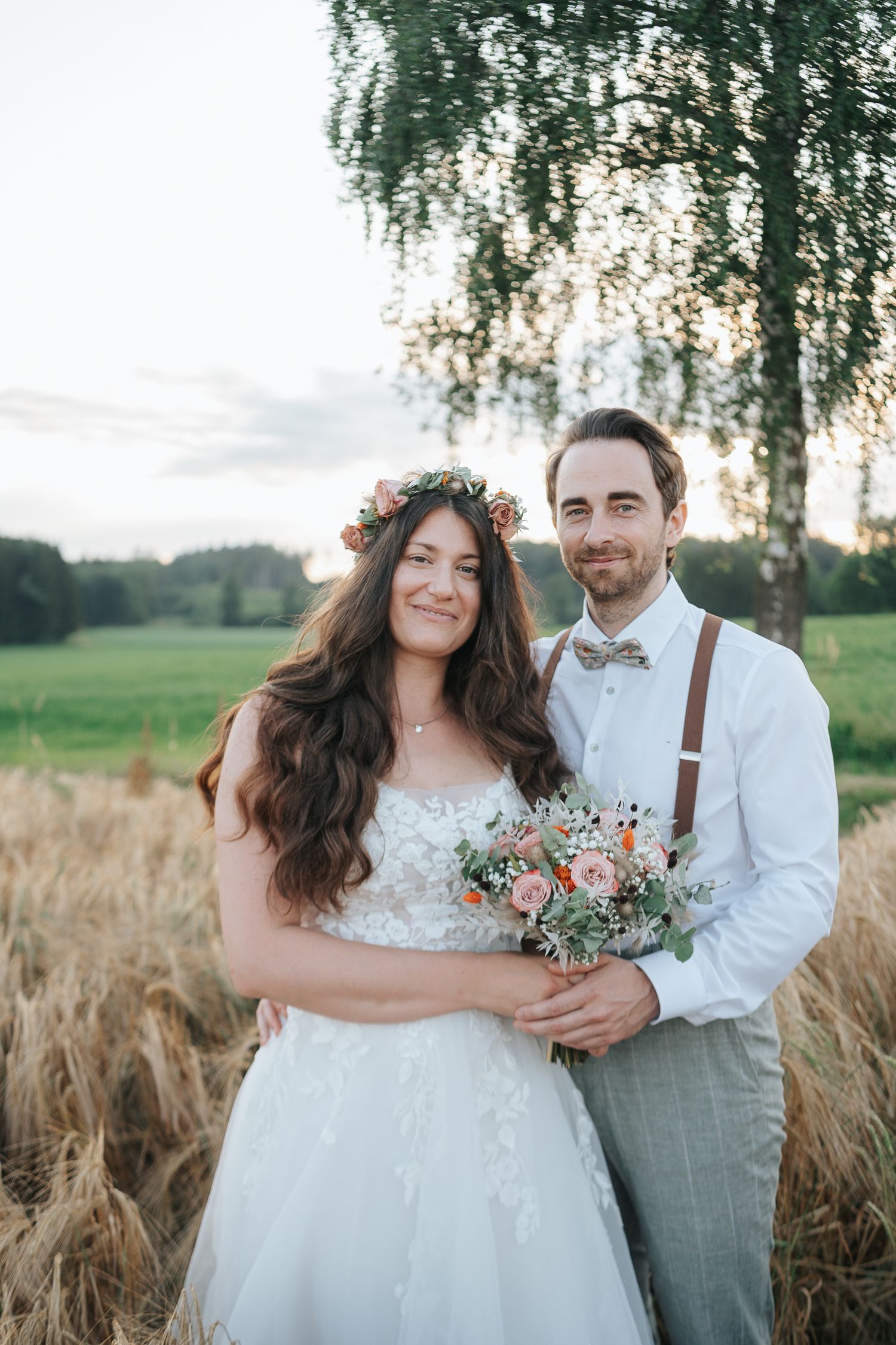 A wedding couple poses in a wheat field at sunset, bride wearing a flower crown and holding wildflowers.