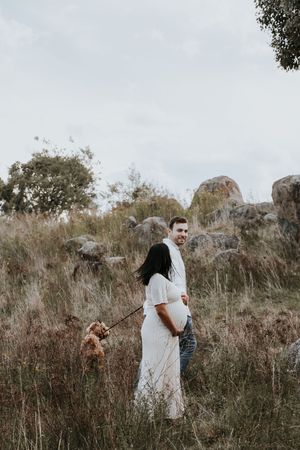 Pregnant couple walking a dog in a field with rocks and trees, woman in white dress, man in jeans smiling.
