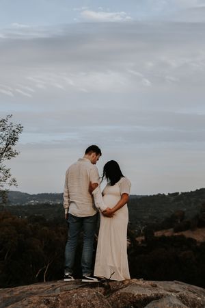 Couple standing on a rock overlooking a scenic landscape during a maternity photoshoot at golden hour.