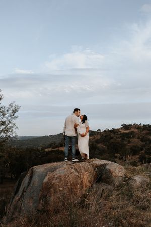 Couple sharing a romantic kiss on a boulder with scenic rolling hills and cloudy sky backdrop.