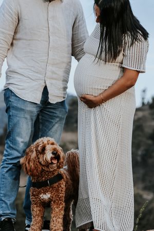 Pregnant woman in white crochet dress stands with partner and fluffy golden doodle dog outdoors.