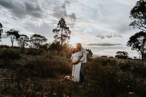 Couple embracing at golden hour in a wildflower field with eucalyptus trees and dramatic cloudy sky.