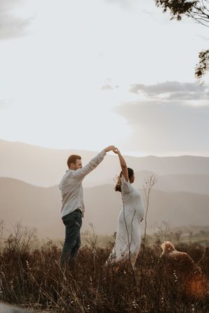 Couple dancing together at sunset with rolling mountains in the background, man spinning woman in white dress.