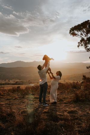 Family lifting child at golden sunset in mountain landscape, warm light glowing behind them on a grassy hilltop.