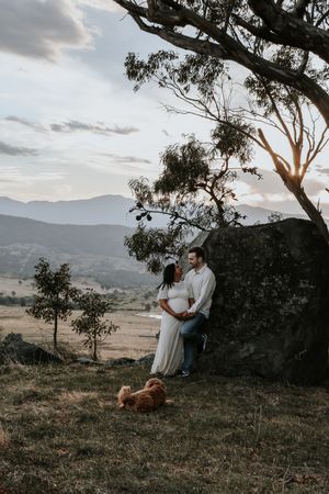 Pregnant couple embracing at sunset by a boulder with mountain views and a small dog nearby.
