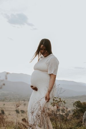 Pregnant woman in white knit dress cradling her baby bump outdoors with mountain landscape backdrop.