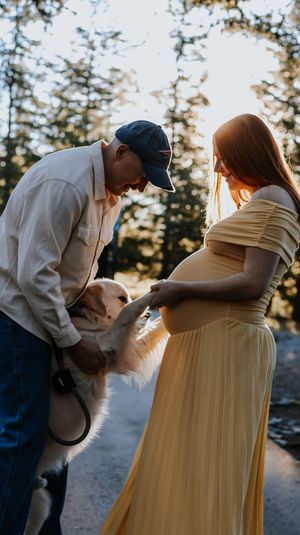 Pregnant woman in yellow dress and man in cap interact with a golden dog outdoors at sunset.