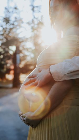 Pregnant woman cradling her baby bump outdoors with golden sunlight streaming through trees.