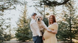 Pregnant woman in yellow dress poses with partner holding white Labrador puppy outdoors among evergreen trees.