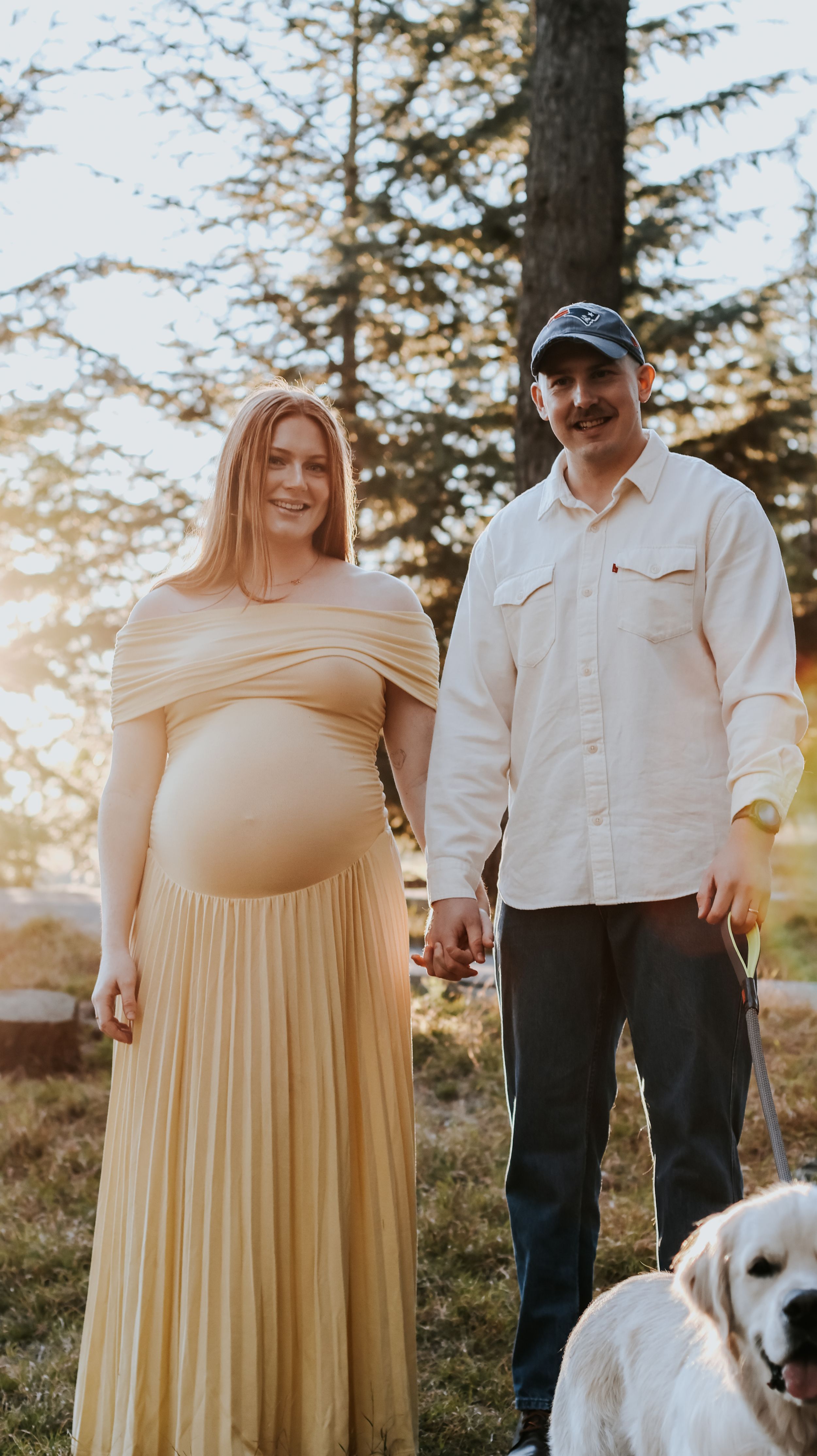 Pregnant woman in yellow dress holds hands with man in white shirt and cap, standing outdoors with a golden retriever.