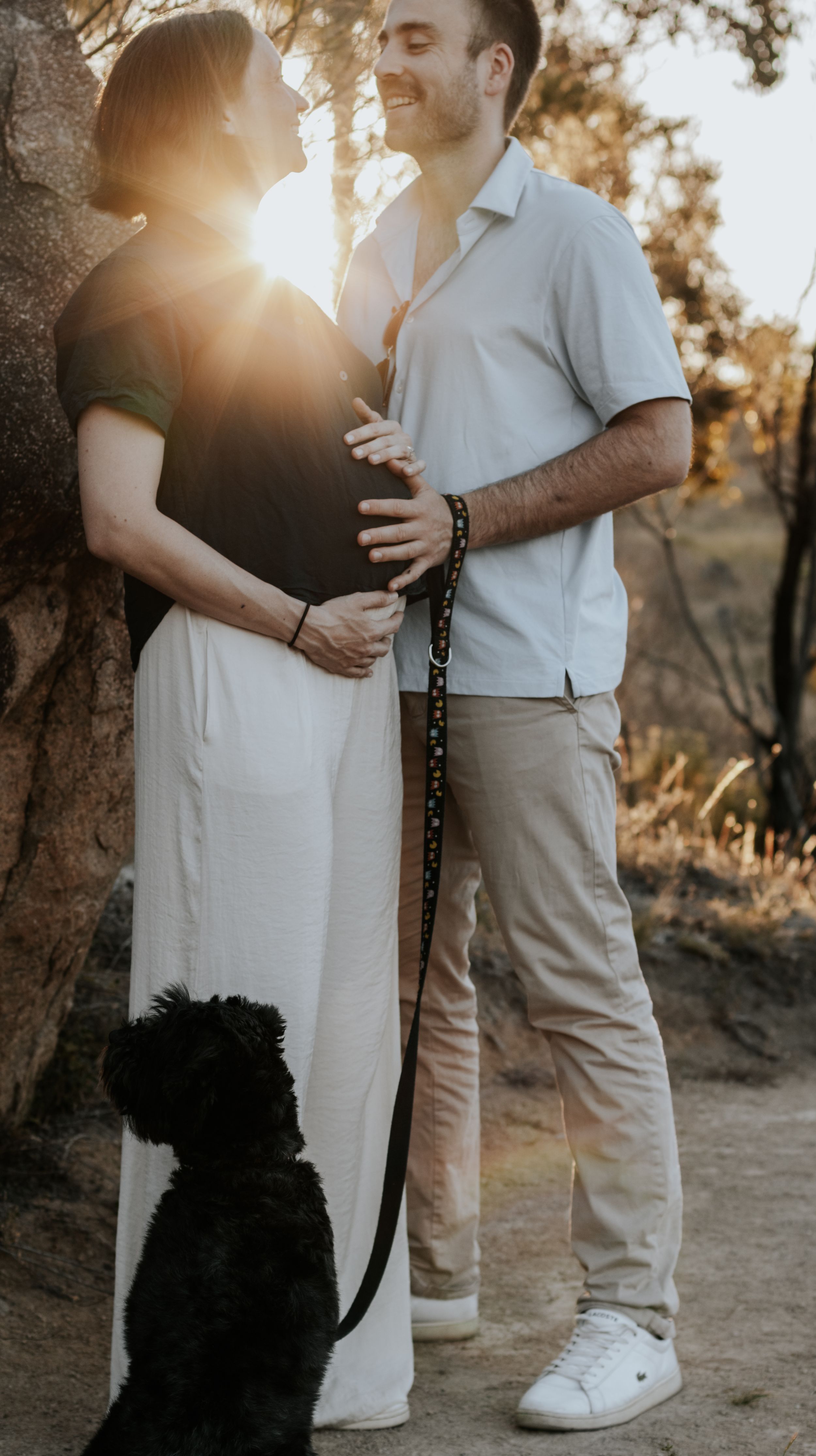 Pregnant couple holding baby bump at golden hour sunset outdoors with black dog on leash beside them.