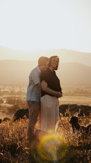 Couple embracing at golden sunset in a field, woman laughing joyfully, dog nearby, mountains in background.