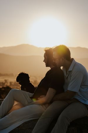 Two men and a dog sit together watching a golden sunset over mountains, silhouetted against warm glowing light.