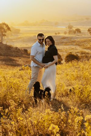 Couple embracing at golden sunset in a field, woman in maternity dress with two black dogs, scenic rural landscape.