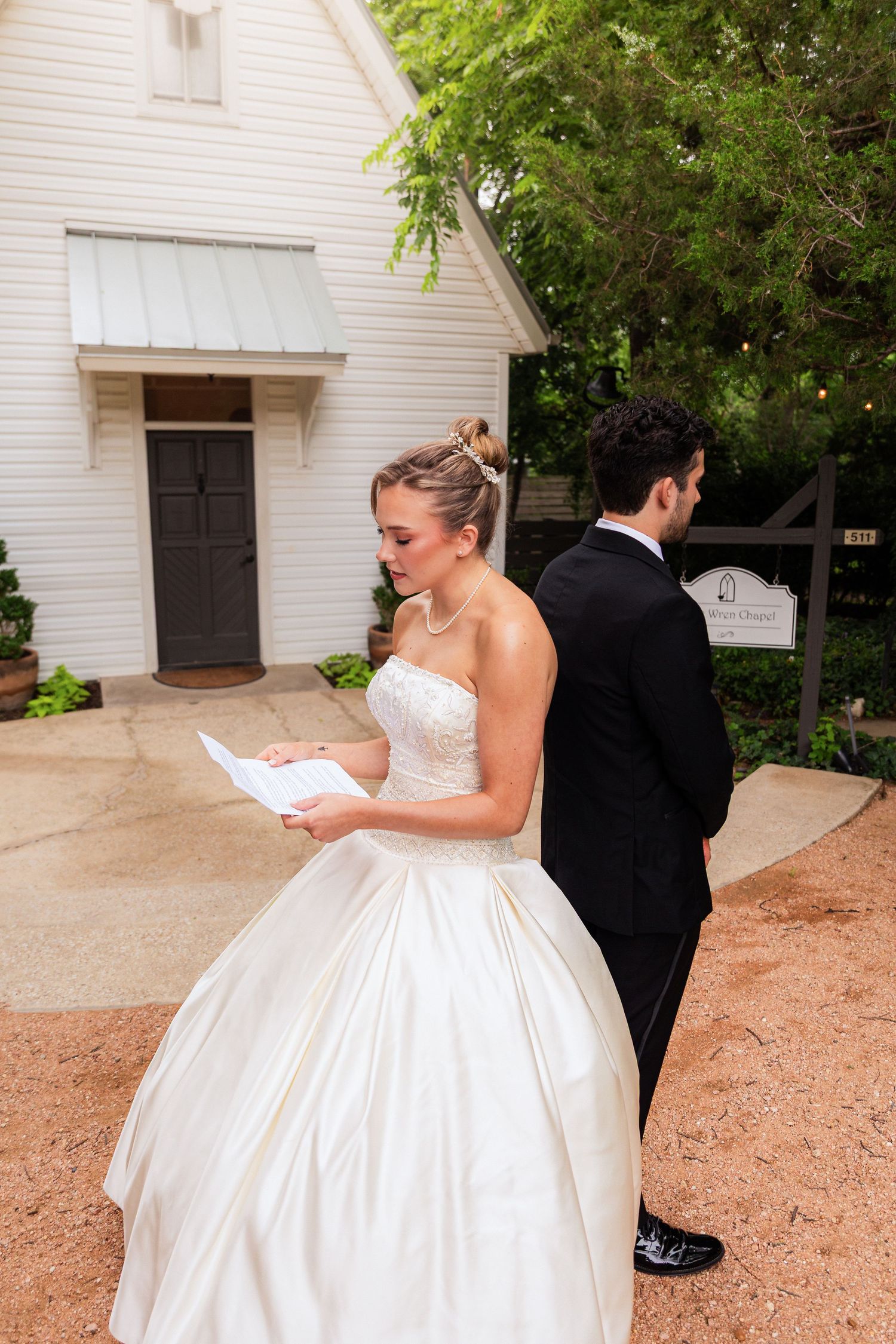 Bride in strapless ball gown reads letter back-to-back with groom in black suit outside white chapel.