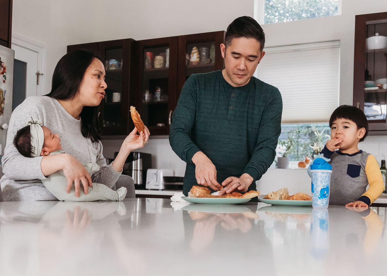Asian family preparing food in kitchen, parents with two young children making sandwiches at white counter.