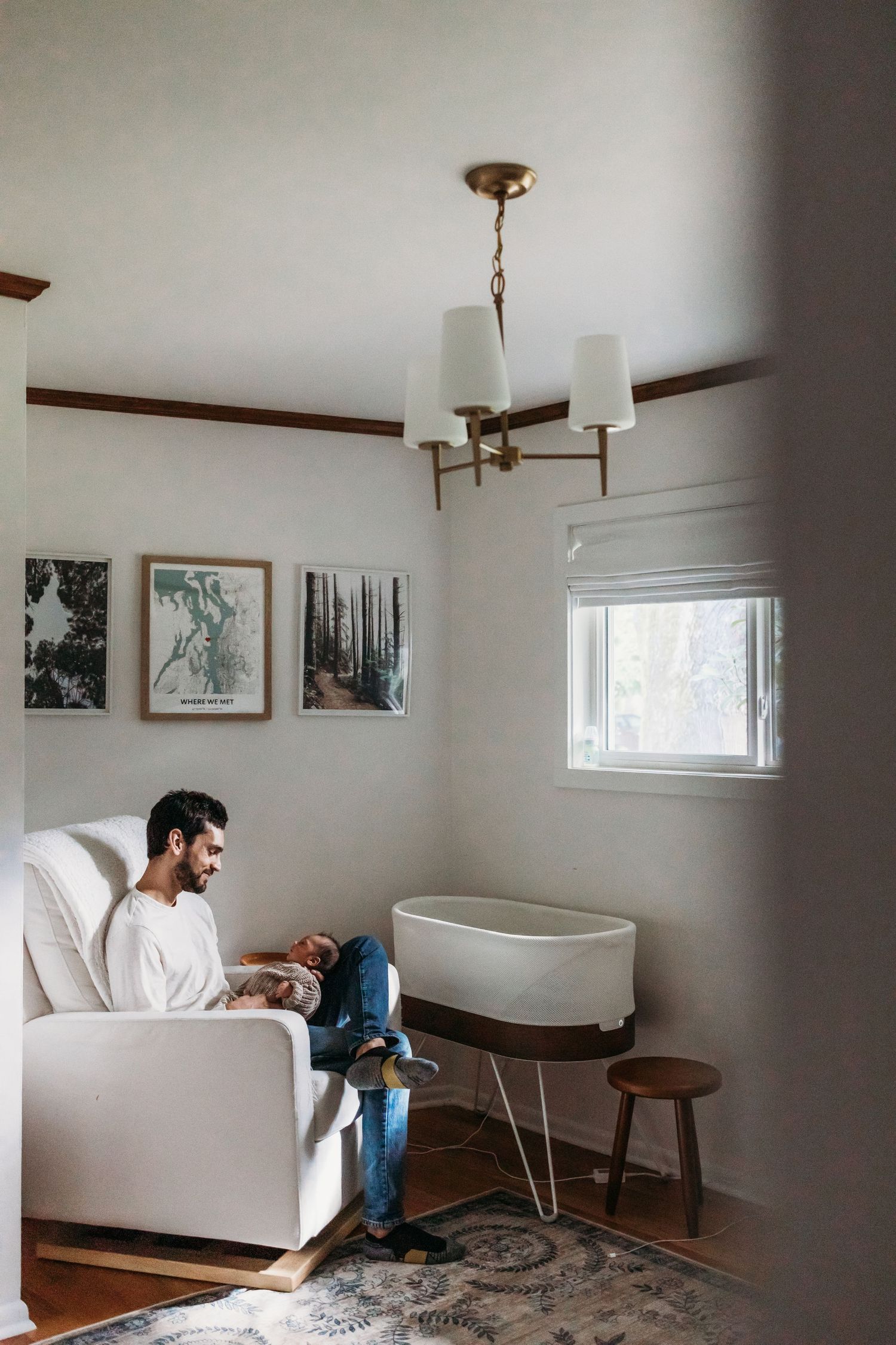Man sitting on white sofa holding baby in cozy nursery with brass chandelier, bassinet, and nature wall art.