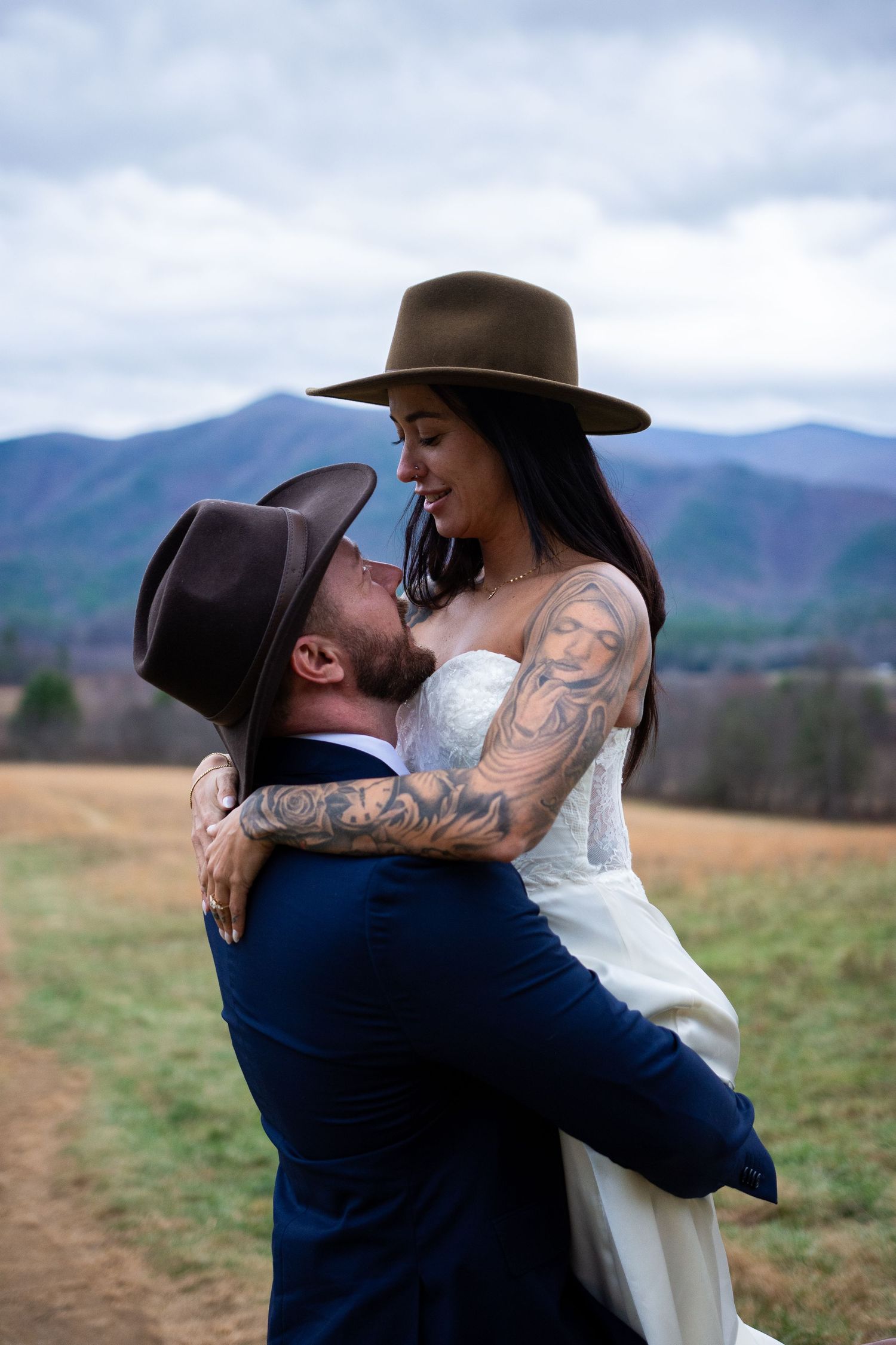 Tattooed bride and groom in wide-brim hats embrace in a mountain field during their outdoor wedding ceremony.