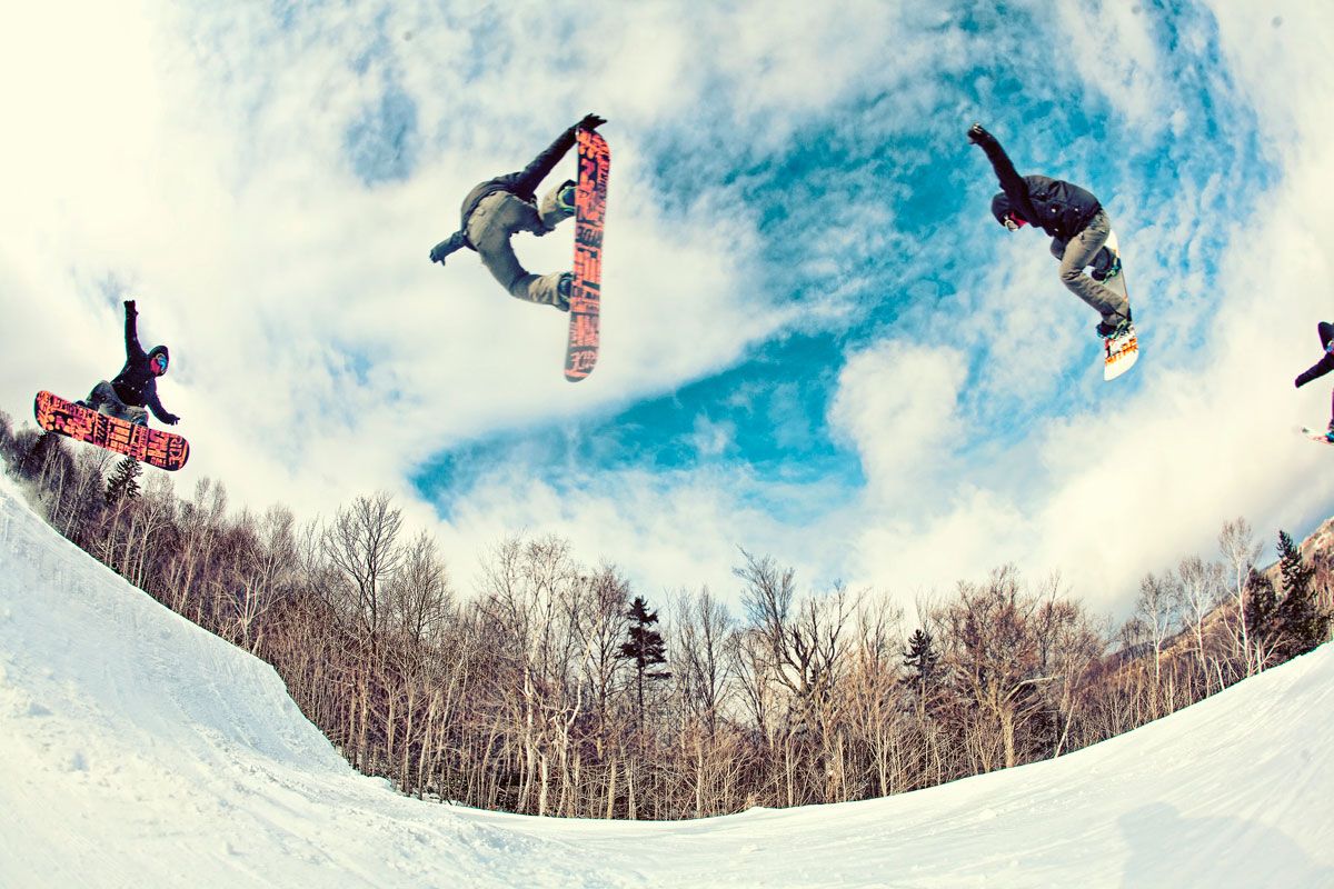 A snowboarder performs multiple aerial tricks in a half-pipe against a blue sky in a snowy winter landscape.