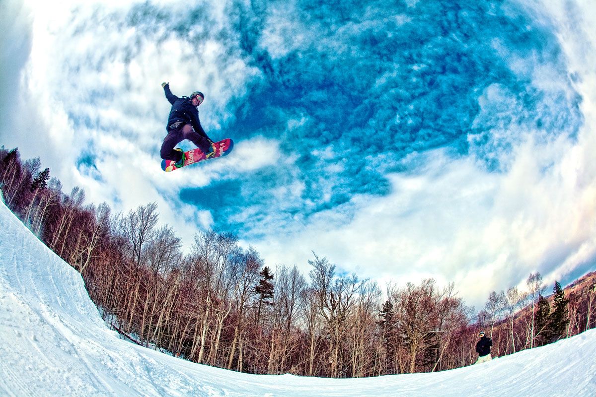 Snowboarder performing a high aerial trick over a snowy halfpipe with bare trees and dramatic cloudy sky.