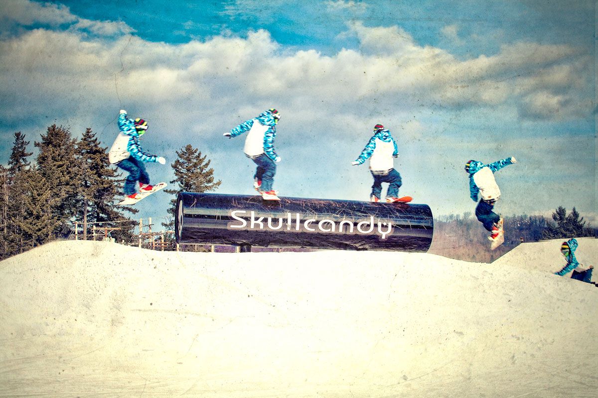 Snowboarders performing tricks on a Skullcandy rail at a snow park under a cloudy blue sky.