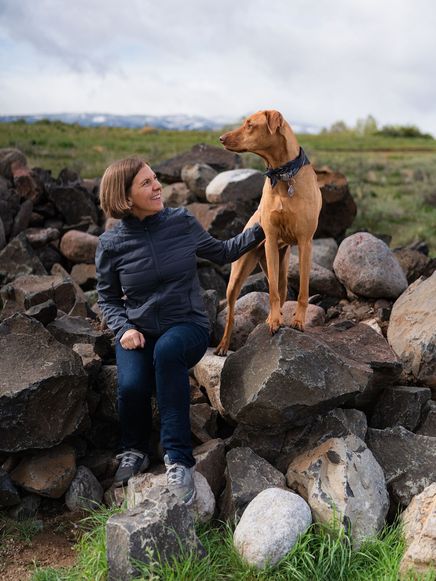 Woman sitting on rocky wall petting a standing Vizsla dog outdoors with green fields and mountains in background.